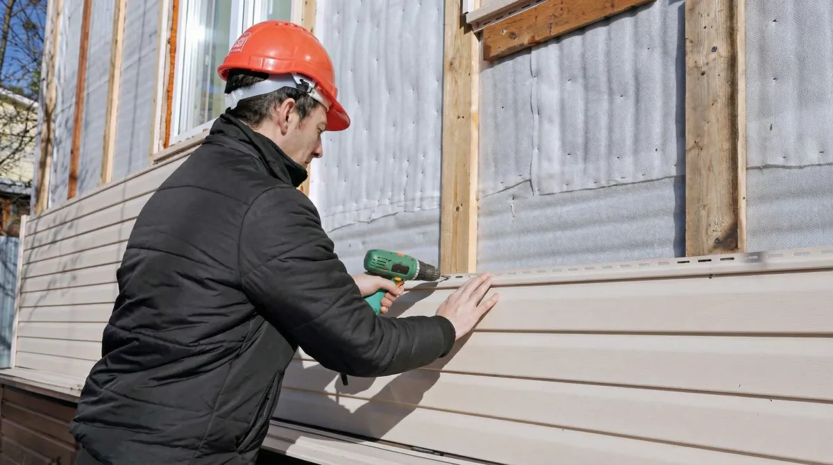 Siding replacement professional using a power drill to install new tan vinyl siding on a home exterior in Overland Park, KS